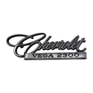 May include: A silver-colored Chevrolet Vega 2300 emblem. The word "Chevrolet" is in a cursive font above a rectangular bar with the text "VEGA 2300". The emblem is made of metal and has a vintage appearance.