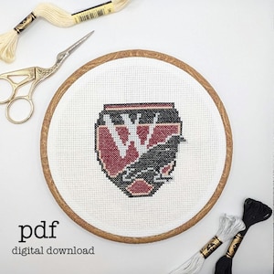 May include: A cross-stitch design featuring a black raven and the letter "W" in a burgundy shield. The design is set in a wooden embroidery hoop. The words "pdf digital download" are at the bottom. Embroidery floss and scissors are also visible.