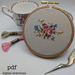 May include: A cross-stitch embroidery hoop with a floral design in pink, blue, and yellow. The wooden hoop is set against a backdrop of a floral teacup, embroidery floss, and gold scissors. The text "pdf digital download" is at the bottom.