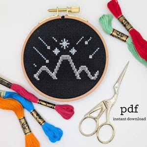 May include: A round wooden embroidery hoop containing black fabric with a cross-stitch design of white and light blue mountains and stars. Surrounding the hoop are several colorful embroidery floss skeins in red, green, pink, blue, and orange, along with a pair of gold-toned stork-shaped embroidery scissors.