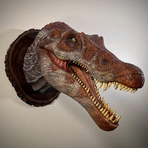 May include: A detailed dinosaur head sculpture in shades of brown and grey, with an open mouth displaying yellow teeth. The sculpture is mounted on a dark brown, circular wooden plaque, suitable for wall display.