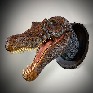 May include: A wall-mounted dinosaur head sculpture featuring textured brown and grey scales, a wide-open mouth with sharp yellow teeth, and a single visible green eye. The creature is attached to a circular, dark brown wood-grain plaque.