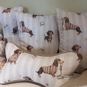 Op de afbeelding: Een collectie decoratieve kussens met een herhalend patroon van bruine en zwarte teckels op een lichtbeige en wit gestreepte achtergrond. De kussens omvatten vierkante vormen en één botvormig kussen met een bruine, getextureerde rand.