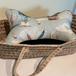 Op de afbeelding: Een decoratief kussen met een lichtblauwe achtergrond met illustraties van boeken, veren en drankflessen. Het kussen ligt in een gevlochten mand met een handvat. Het kussen heeft een zwarte rand.