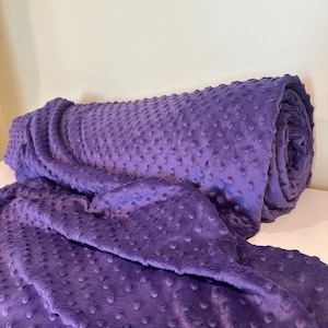 Könnte beinhalten: Eine tiefviolette Decke mit einer erhabenen Noppenstruktur auf der gesamten Oberfläche. Der Stoff ist auf einer Seite zylindrisch aufgerollt, während der restliche Teil der Decke nach vorne drapiert ist und das gleichmäßige Punktmuster zeigt.