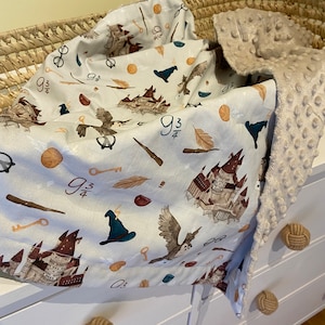 Peut inclure: Une couverture pour bébé avec un fond bleu clair présentant un motif répété d'images de Poudlard, notamment des châteaux, des hiboux, des clés et le chiffre 9 3/4. La couverture est pliée dans un panier en osier, avec un envers doux et beige texturé.