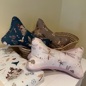 Puede incluir: Cojines decorativos en forma de hueso con varios diseños, incluyendo llaves, castillos y botellas de pociones. Los cojines están en una cesta y sobre una superficie blanca. Los cojines tienen un ribete lila claro y son de tela suave.