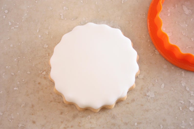 A white, round cookie cutter is placed on a speckled surface next to an orange cookie cutter.
