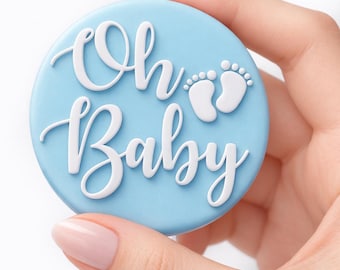 Może przedstawiać: Jasnoniebieska, okrągła dekoracja na baby shower z napisem "Oh Baby" w białym skrypcie. Dwa białe odciski stóp dziecka znajdują się nad słowem "Baby".