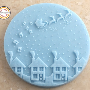 Puede incluir: Un sello para galletas de silicona azul con una escena de invierno. El sello representa un paisaje nevado con casas, una chimenea y Papá Noel volando en un trineo tirado por renos. El sello es redondo y tiene un diámetro de aproximadamente 7,5 cm.