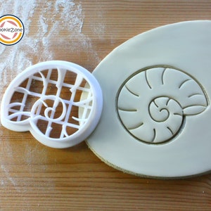 May include: A white plastic cookie cutter shaped like a seashell, with a detailed design. The cutter is shown on a white fondant surface, with a seashell imprint.