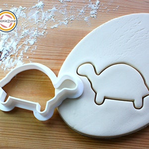 May include: White plastic turtle cookie cutter on a wooden surface with a white fondant turtle cutout. The cookie cutter is on the left and the fondant cutout is on the right.