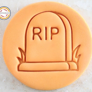 Może przedstawiać: Pomarańczowa, okrągła foremka do ciastek z motywem nagrobka. Na nagrobku widnieje napis "RIP", a po bokach trawa. Idealna do tworzenia ciasteczek na Halloween.