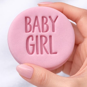 以下が含まれることがあります： 表面に「BABY GIRL」の文字がエンボス加工された、丸い薄いピンク色の物体。テキストはサンセリフ体です。ピンクのマニキュアを塗った手で持っています。