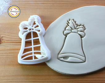 May include: White plastic Christmas bell cookie cutter with a bow and holly leaves on top. The cutter is shown next to a white clay impression of the cutter.