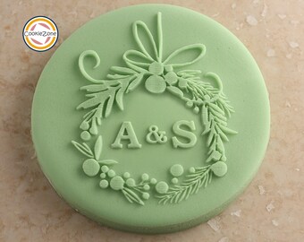 May include: A round, light green cookie with a wreath design and the letters "A & S" in the center. The wreath features leaves, berries, and a bow at the top. The CookieZone logo is in the upper left corner.