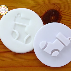 Op de afbeelding: Twee witte, cirkelvormige koekjessnijders. Eén snijder heeft een verhoogd ontwerp van een kerstsok. De andere snijder heeft hetzelfde sokontwerp, maar het ontwerp is in de snijder geperst. Het CookieZone-logo staat in de linkerbovenhoek.