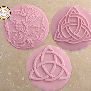 Può includere: Tre timbri per biscotti rosa con disegni in rilievo. Uno presenta un motivo di cardo e pigne, mentre gli altri due mostrano disegni di nodi celtici. I timbri sono circolari e realizzati in un materiale rosa chiaro.