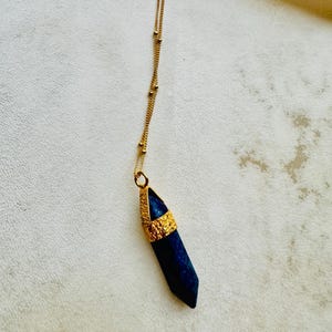 May include: A gold chain necklace with a dark blue crystal pendant. The pendant is a pointed, faceted crystal with a gold setting.