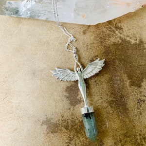 Pode incluir: Colar de prata com um pingente que apresenta uma figura de anjo detalhada segurando uma ponta de cristal verde e preta.