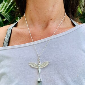 Pode incluir: Um colar de prata com um pingente que apresenta uma figura feminina alada em p&eacute; sobre um cristal verde e cinza.