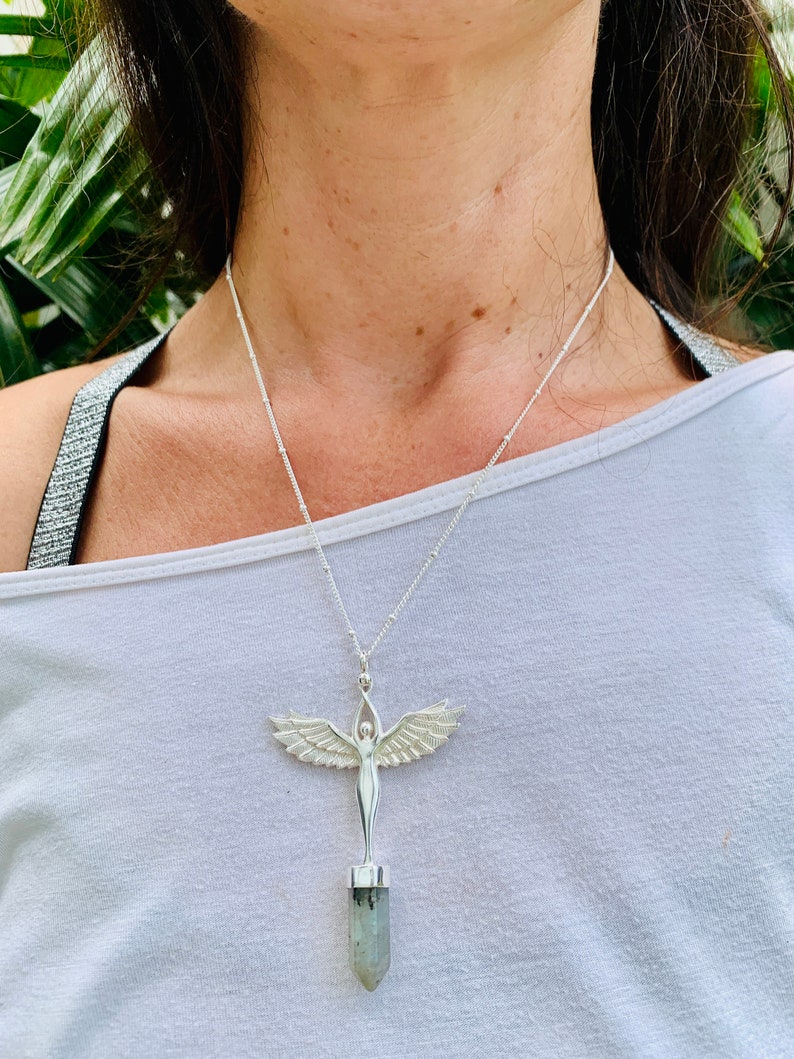 Pode incluir: Um colar de prata com um pingente que apresenta uma figura feminina alada em p&eacute; sobre um cristal verde e cinza.