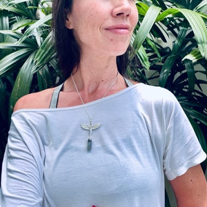 Pode incluir: Um colar de prata com um pingente de cristal e um design de asa de anjo. O colar &eacute; usado por uma pessoa com cabelo castanho comprido e uma camisa branca.