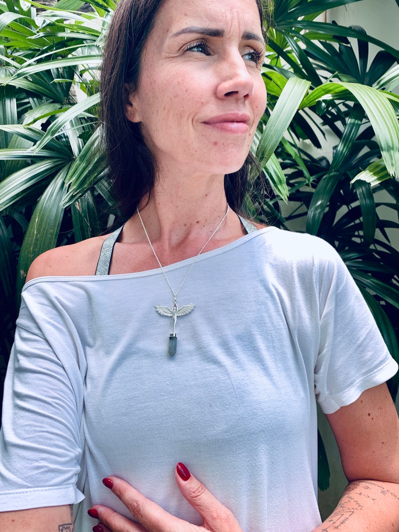 Pode incluir: Um colar de prata com um pingente de cristal e um design de asa de anjo. O colar &eacute; usado por uma pessoa com cabelo castanho comprido e uma camisa branca.