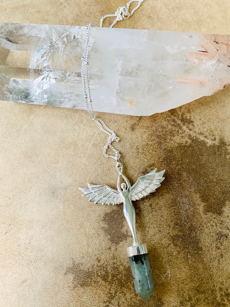 Pode incluir: Colar de prata com um pingente que apresenta uma figura de anjo detalhada segurando uma ponta de cristal verde e preta.