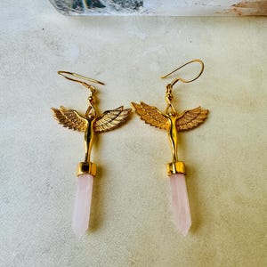 Può includere: Un paio di orecchini dorati con ali d'angelo e pendenti in cristallo di quarzo rosa. Gli orecchini presentano un design dettagliato di angelo con ali spiegate e chiusura a gancio. I cristalli di quarzo sono di colore rosa pallido.
