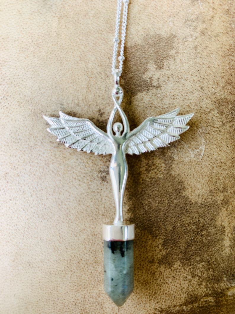 Pode incluir: Colar de prata com um pingente que apresenta uma figura de anjo detalhada com asas e uma pedra preciosa cinza na parte inferior.