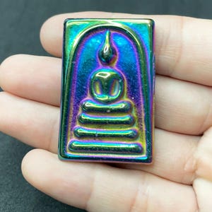 Könnte beinhalten: Ein kleiner, rechteckiger, schillernder Amulet mit einem erhabenen Bild eines sitzenden Buddha. Der Amulet ist aus Metall und hat eine regenbogenartige Oberfläche.