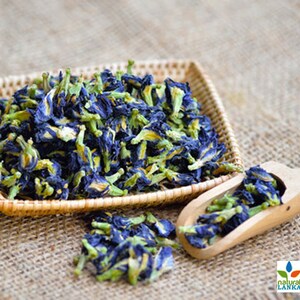 May include: Dried blue butterfly pea flowers in a woven basket and a wooden scoop. The flowers are a vibrant blue with yellow centers and green stems.