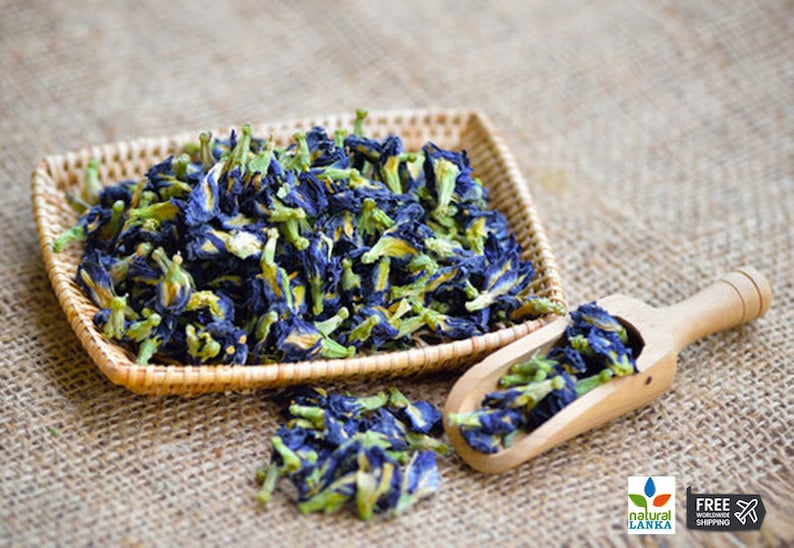 May include: Dried blue butterfly pea flowers in a woven basket and a wooden scoop. The flowers are a vibrant blue with yellow centers and green stems.