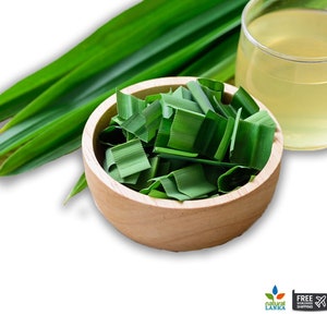 May include: A wooden bowl filled with chopped green pandan leaves, accompanied by several long, whole pandan leaves and a clear glass containing a pale yellow liquid. The composition features fresh botanical elements arranged on a white background.