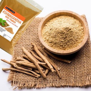 Op de afbeelding: Een bruine zak met Ashwagandha-wortelpoeder met de tekst "Natural Lanka Ashwagandha Root Powder (Withania somnifera)" op het etiket. Een kom met het poeder staat op een jute oppervlak met gedroogde wortelstukken eromheen.