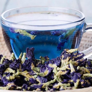 May include: A glass cup filled with blue butterfly pea flower tea, surrounded by dried blue butterfly pea flowers.