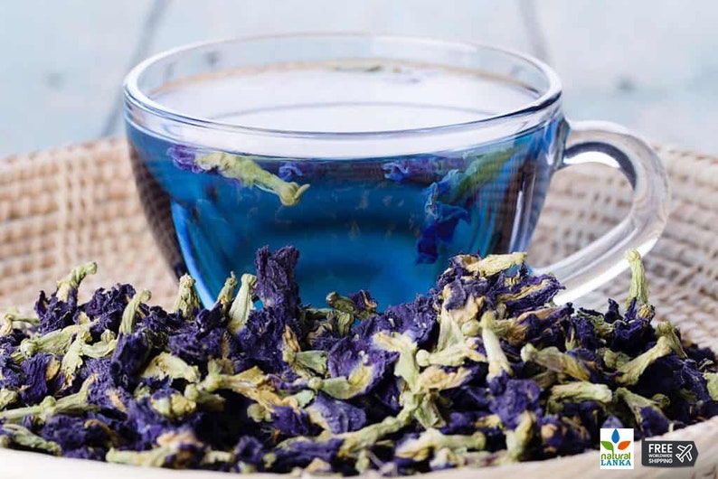 May include: A glass cup filled with blue butterfly pea flower tea, surrounded by dried blue butterfly pea flowers.