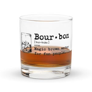 May include: A clear glass tumbler filled with amber liquid, labeled "Bourbon" with a definition and the phrase "Magic brown water for fun people." The glass has a classic, rounded shape and is partially filled.