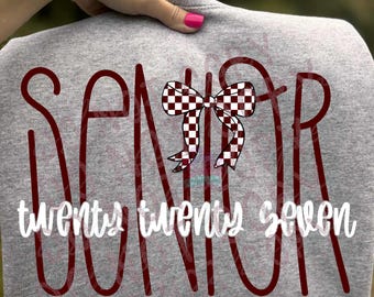 Puede incluir: Una camiseta gris jaspeado con el texto rojo oscuro SENIOR en una fuente alta y delgada. Un gráfico de lazo a cuadros blanco y negro reemplaza la letra I. Debajo, el texto blanco twenty twenty seven está impreso en letra cursiva.