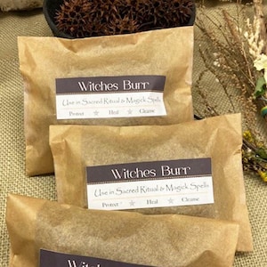 May include: Three brown paper packages labeled "Witches Burr" with text indicating use in sacred rituals and magick spells. The packages are arranged in front of a basket filled with brown seed pods.