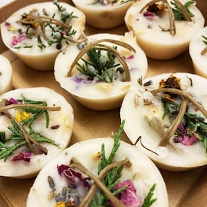 May include: A collection of round, off-white wax melts arranged in a brown tray. Each melt is embedded with dried green cedar sprigs, small purple and yellow flower petals, and thin, curved brown decorative twigs across the top surface.