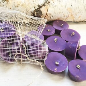 May include: A selection of small, round, purple tea light candles with a natural wick. Some candles are loose, others in a mesh bag. The background features birch bark and a white wooden surface.