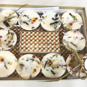 May include: A set of nine white round candles with various dried botanicals and red berries embedded in the wax. The candles are arranged in a box with a plaid pattern and a tag that reads "Aflare Firestarters".