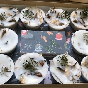May include: A box containing nine white candles, each with natural elements embedded in the wax. The candles are arranged in three rows of three. Each candle features a small sprig of greenery and other natural elements embedded in the wax. The box is lined with patterned paper.