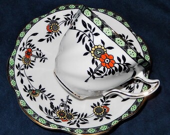 May include: White teacup and saucer set with a floral design. The cup and saucer feature black, orange, and yellow flowers with black leaves. The saucer has a scalloped edge with a green and black geometric border and gold trim.
