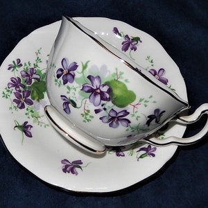 May include: White teacup and saucer set with a floral violet design. The cup and saucer have a gold trim. The violet flowers are purple with green leaves. The teacup has a curved handle.