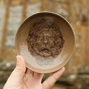 May include: A small, round, brown ceramic dish featuring a detailed, embossed face with a beard. The face is centered in the dish, with intricate details in the hair and facial features. The dish has a slightly raised edge.
