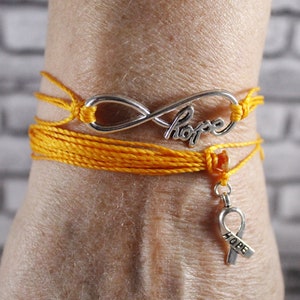 May include: A handmade yellow string bracelet with a silver infinity charm that says "hope". A silver ribbon charm with the word "hope" hangs below. The bracelet is wrapped around a wrist.