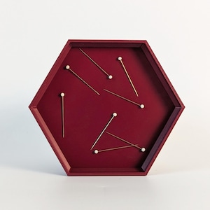 May include: A burgundy, hexagonal tray with several gold-colored pins with white heads. The pins are scattered across the tray's surface. The tray is set against a white background.